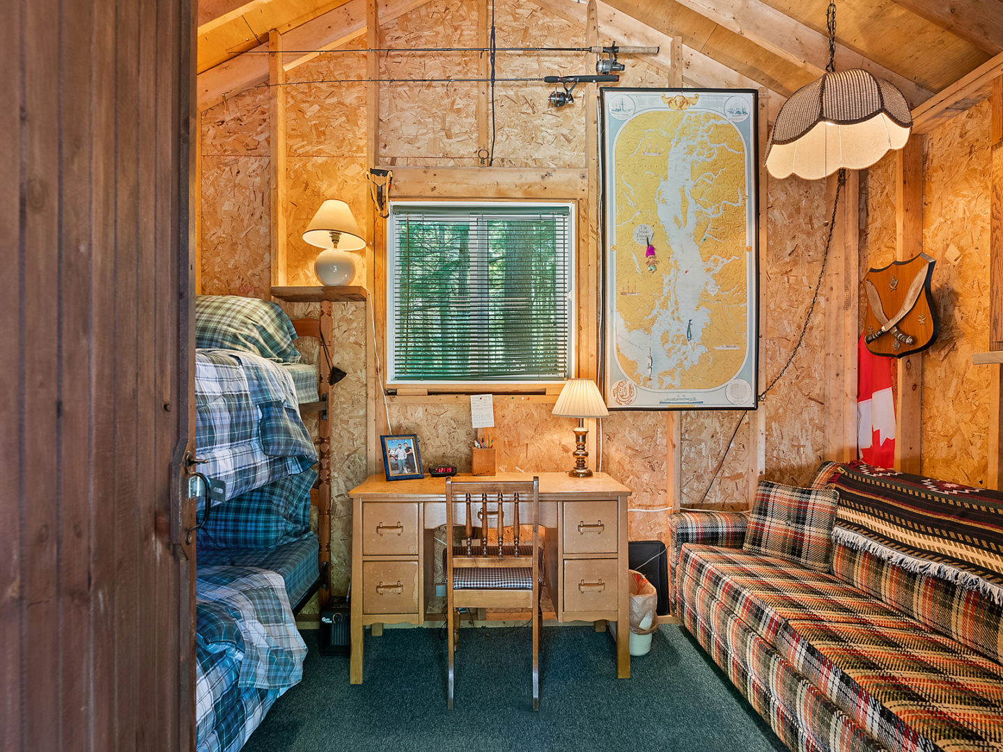 Inside of a small bunkie. A desk sits in the middle of the back wall, and there is a couch to one side and bunkbeds to the other side of it.