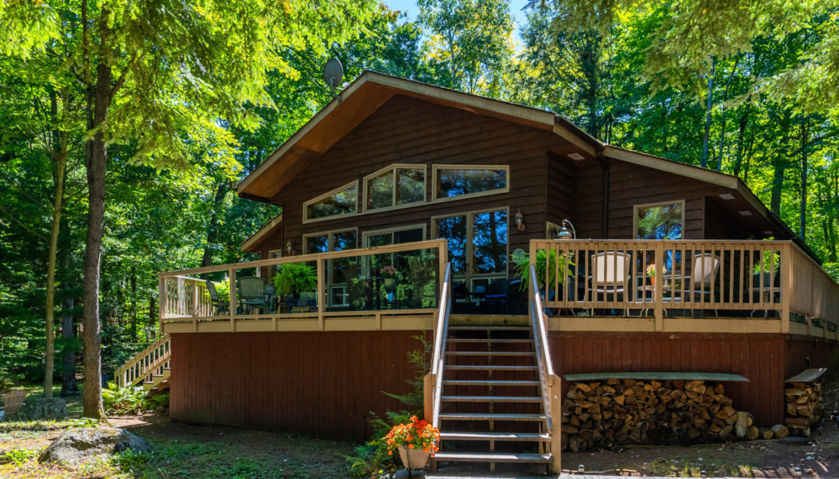 A wood frame cottage with steps leading up to a large deck, which branches off an exterior wall with large windows.