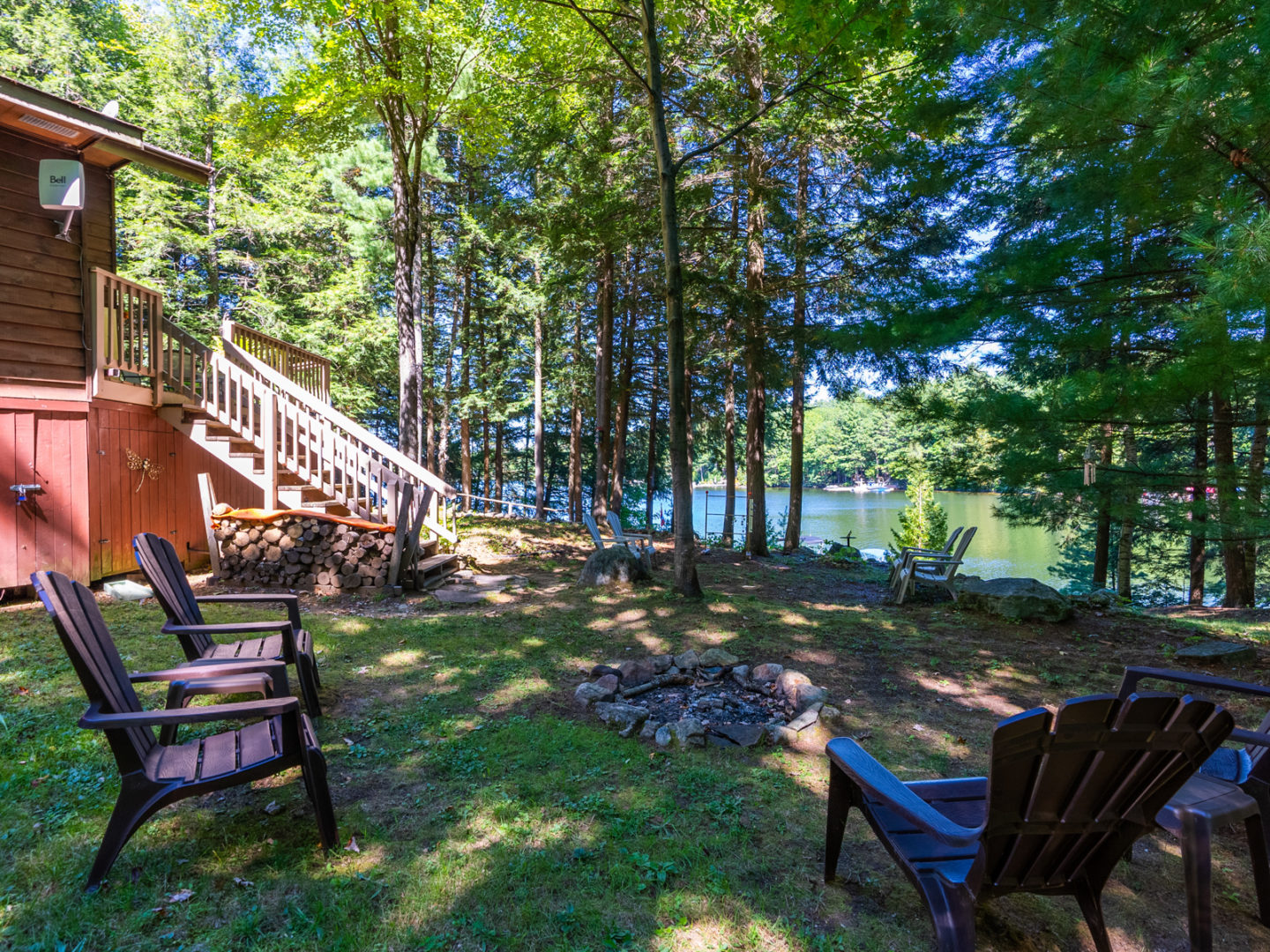 A grassy outdoor area beside a cottage, on a hill by the waterfront. A small fire pit in the middle is surrounded by a few Muskoka chairs.