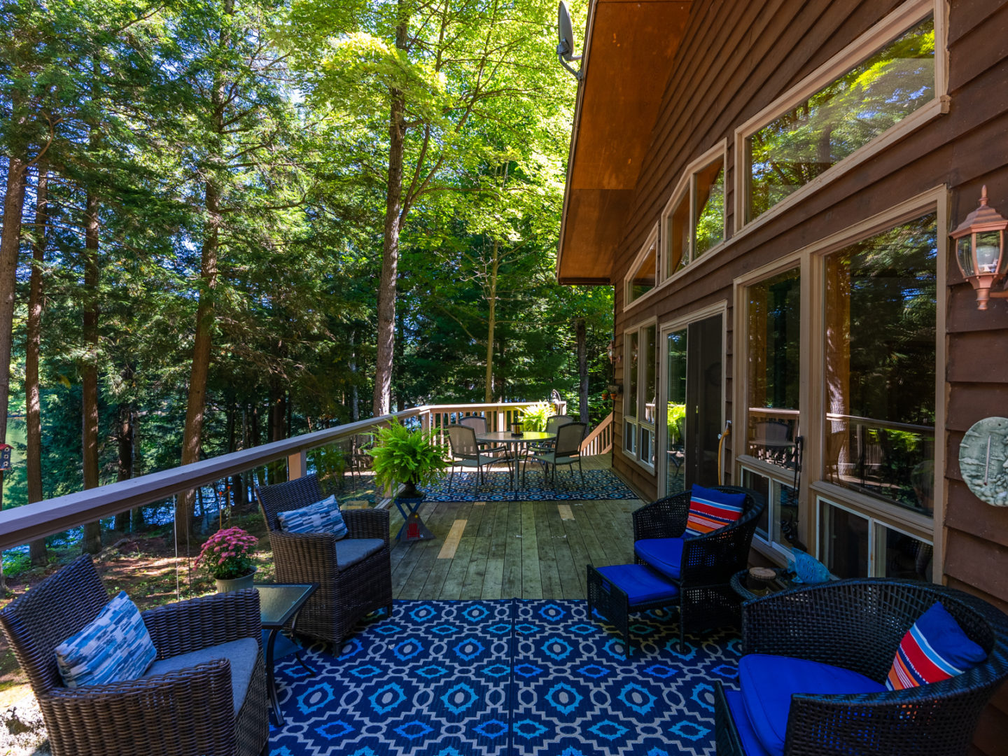 A large deck with an outdoor rug and patio furniture, branching off a cottage with floor-to-ceiling windows.