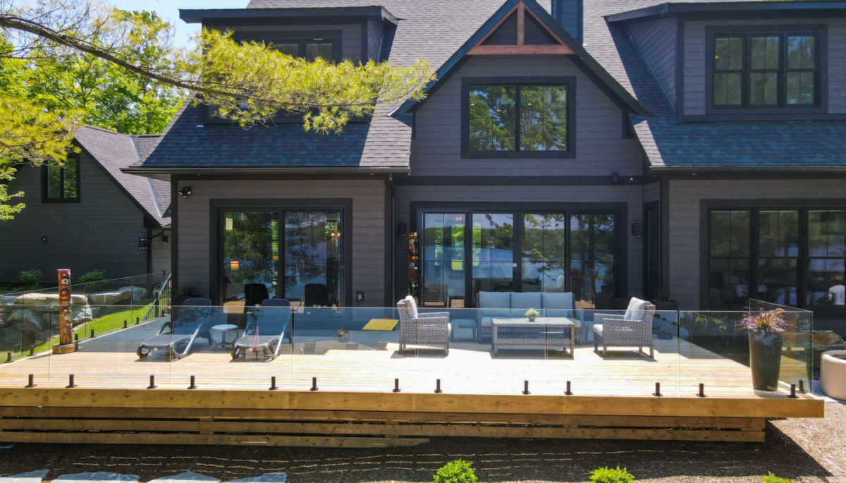 The back of a large, modern cottage with two stories and many wide windows. A large deck with a glass railing branches off the house.