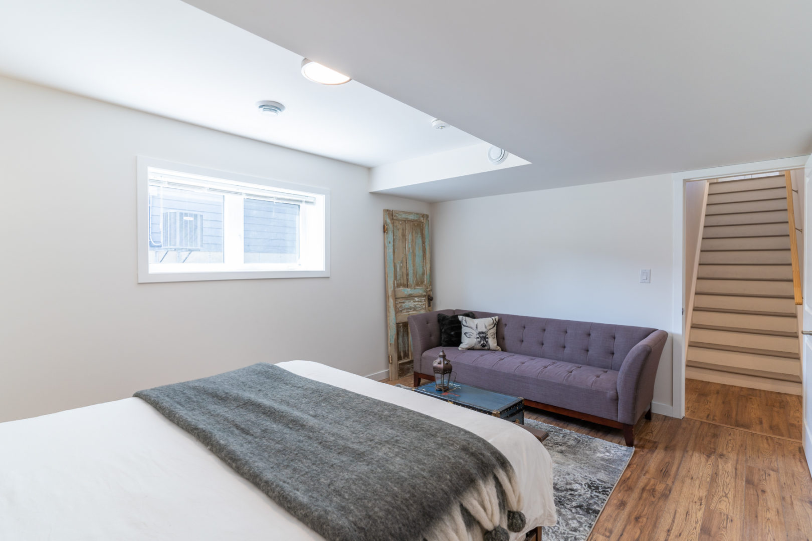 A basement bedroom off the bottom of a staircase, with a big bed, a couch, and a small window.