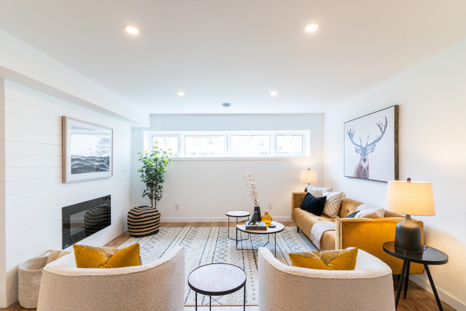 A bright basement living room area with a long window, chairs, a couch, and a fireplace.