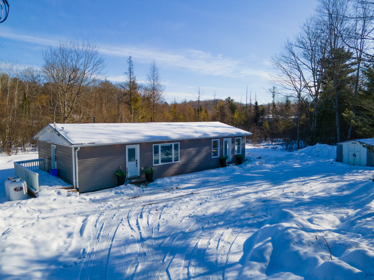 A modest-sized cottage bungalow surrounded by a snowy yard and trees on all sides.