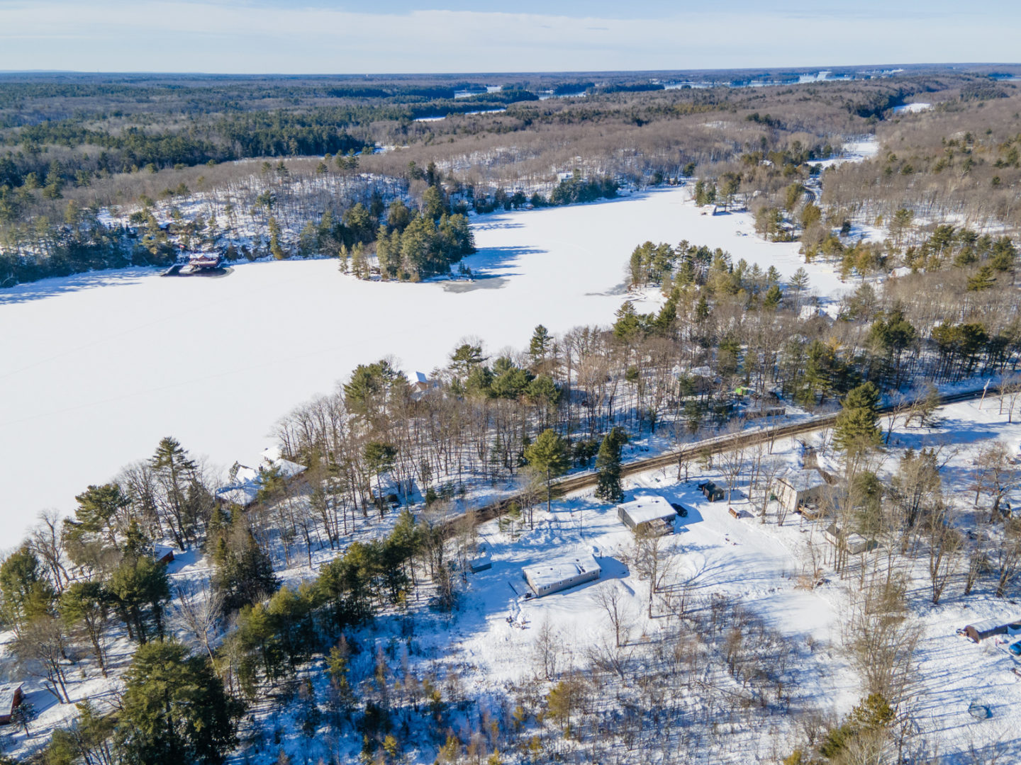 A small stretch of lake in the winter, covered in snow. A small cottage bungalow sits inland, across a road but close to the water.