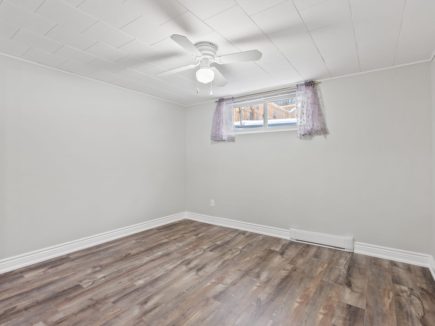 A spacious, empty room with a small window, hardwood floors, and a ceiling fan.