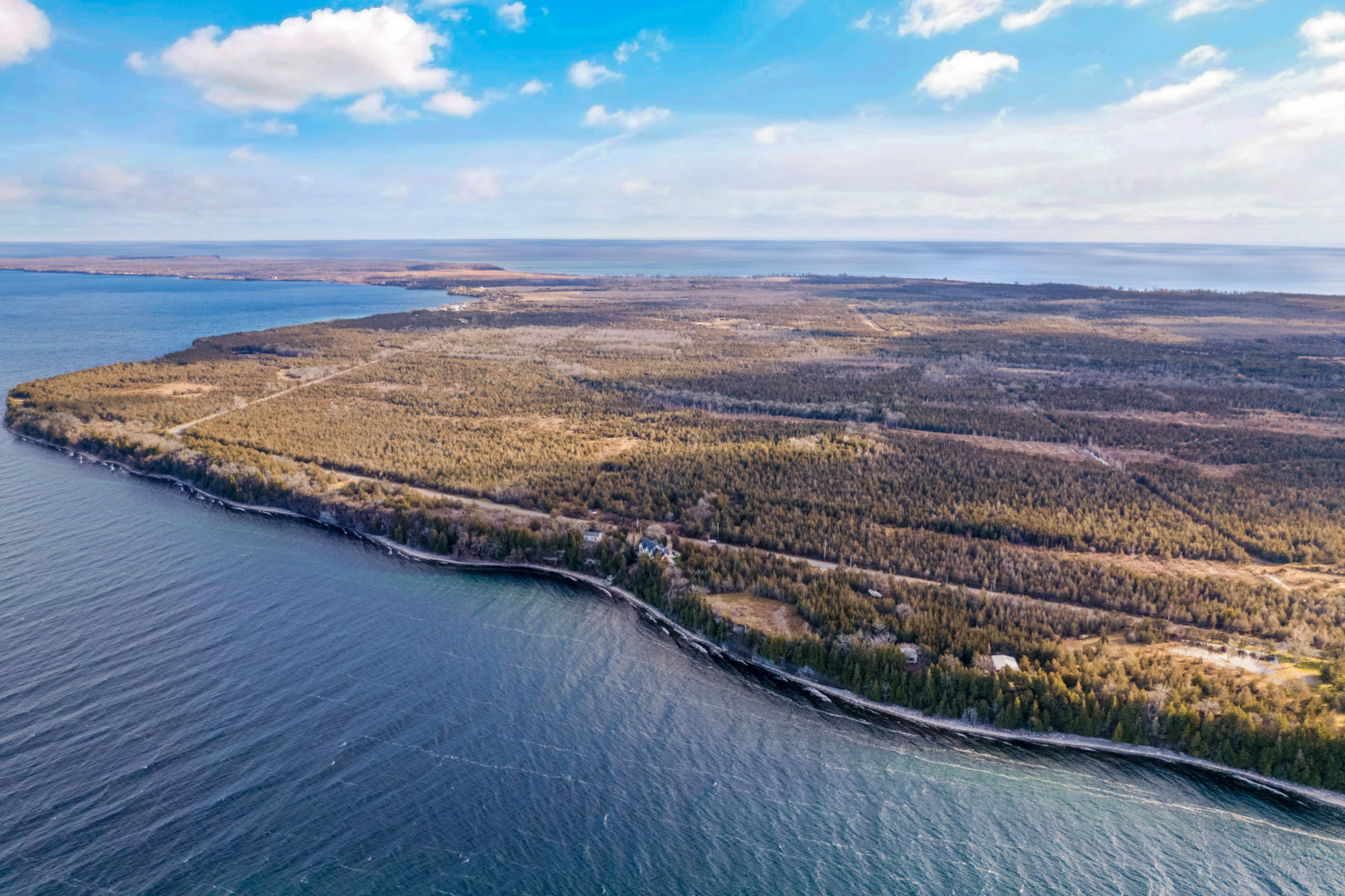 The shoreline of a deep blue lake, filled with a forest of trees. One lone house can be seen along the shore.