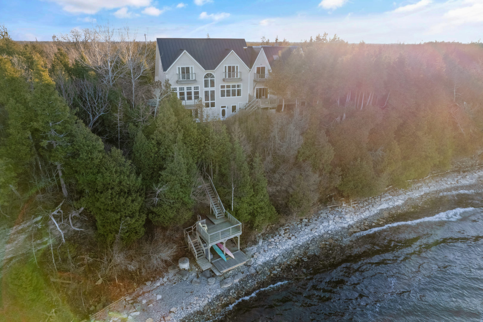 A long, rocky shoreline of a big lake house. A long wooden staircase leads down from the house to the waterfront.
