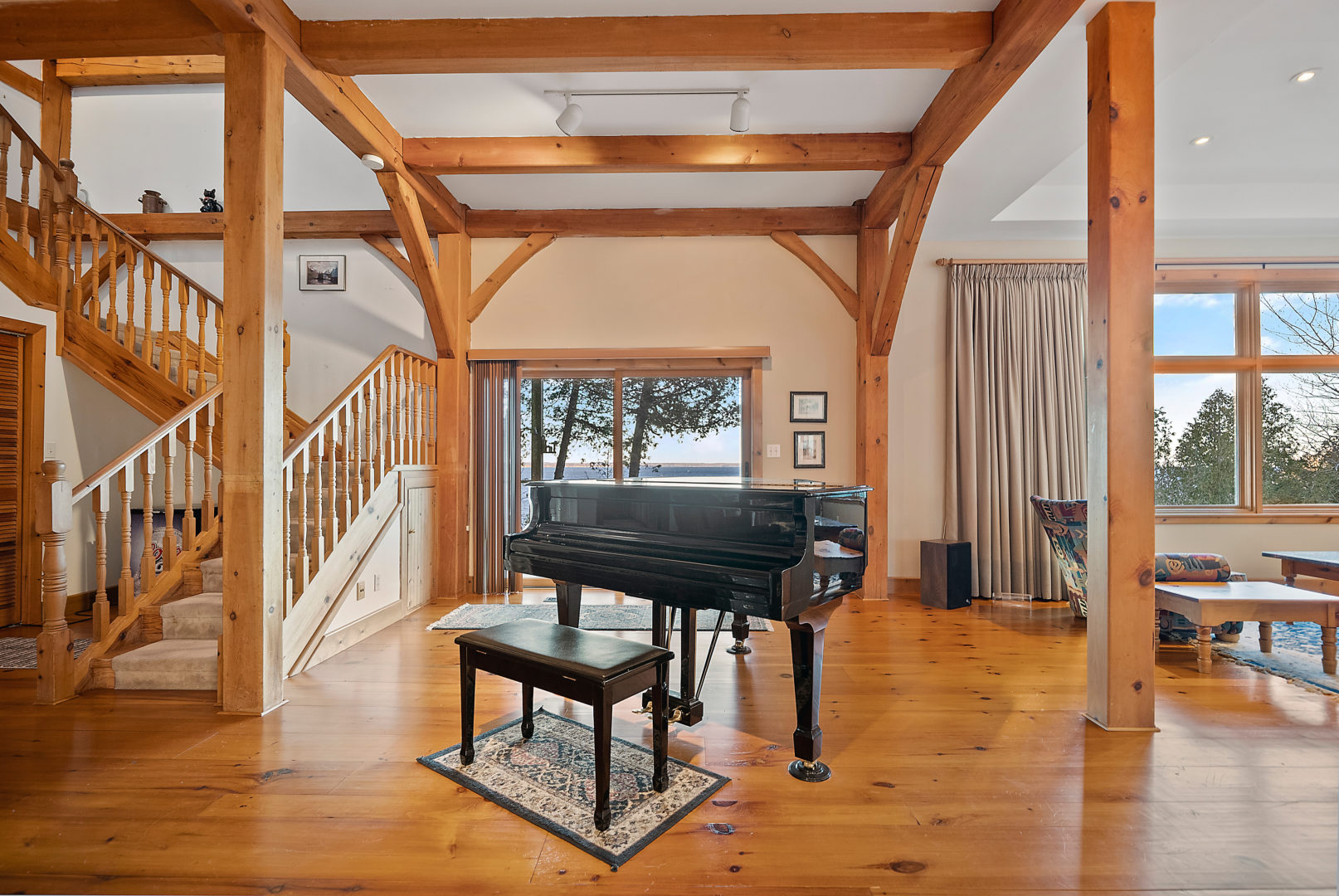 A large grand piano sits in a bright, warm living area, in front of a large window and a staircase.