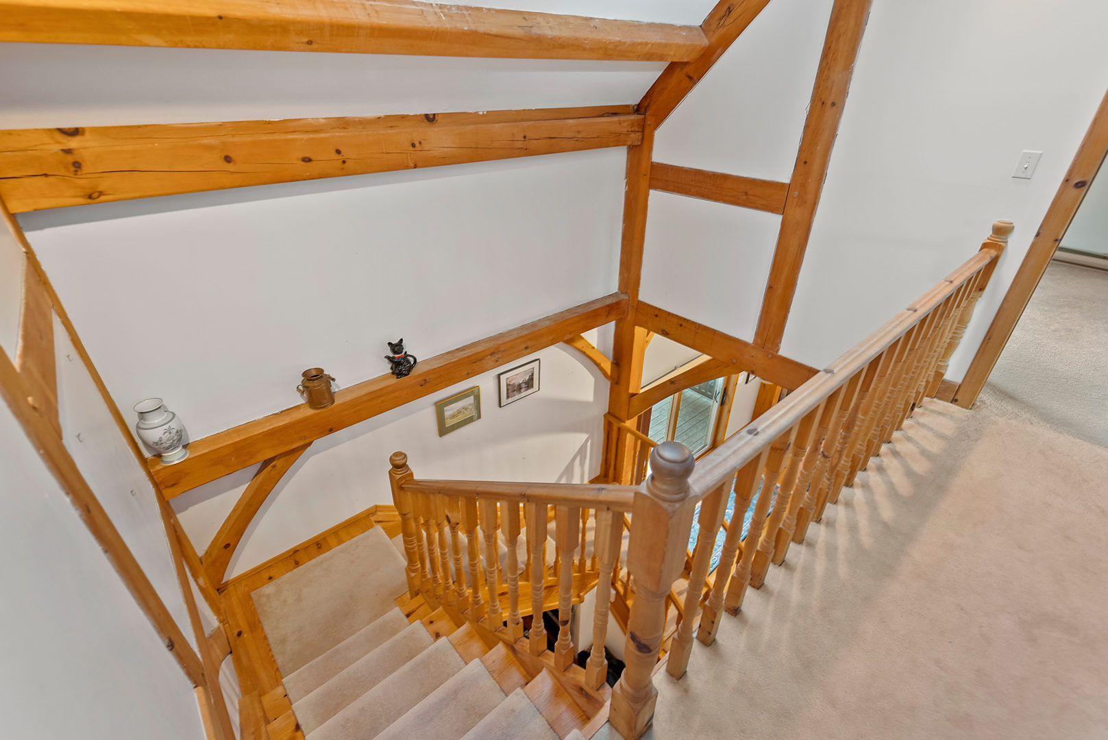 A carpeted staircase leads up to the second story of a large house. A light-coloured wooden railing and wooden beams stand out against white walls and light gray carpeting.
