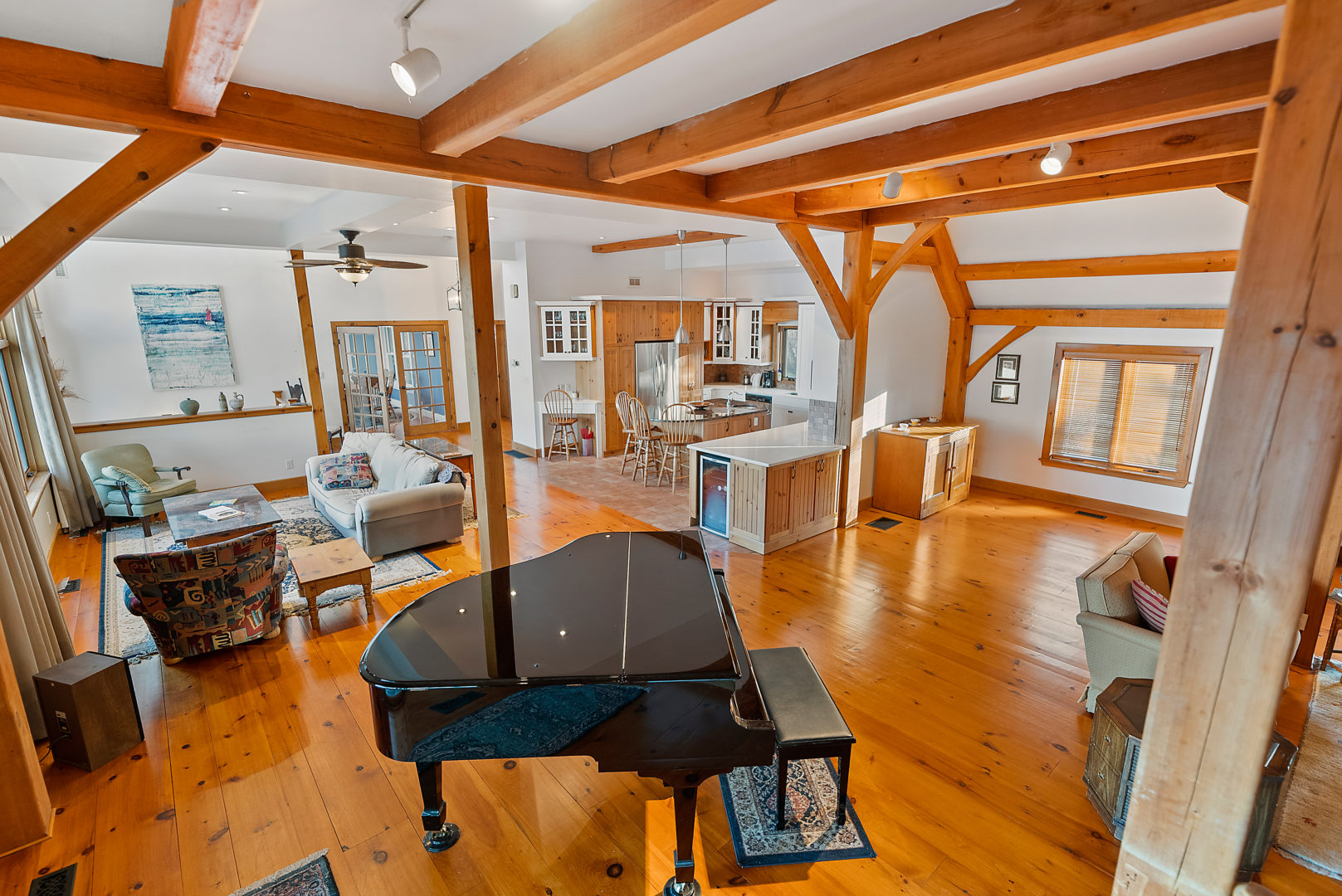 A large open-concept living space with two sitting areas, a grand piano, and a kitchen. Light hardwood floors throughout. Wooden beams extend across a white ceiling, and wooden posts offer support throughout the space.