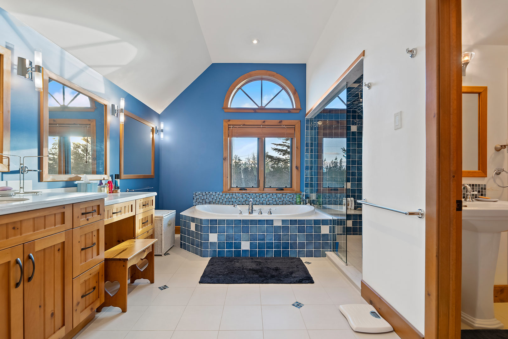 A large bathroom with a bathtub and a shower. Blue tile lines the side exterior of the tub and a painted blue accent wall with a large window sit behind it.