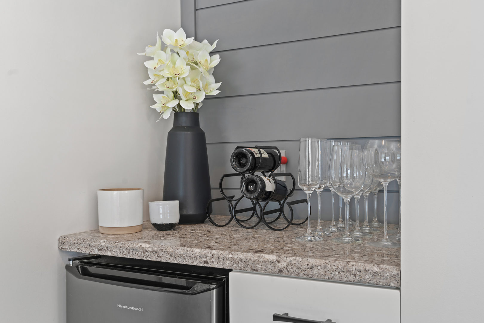 A small bar space with a mini fridge below the counter and wine glasses on the counter.
