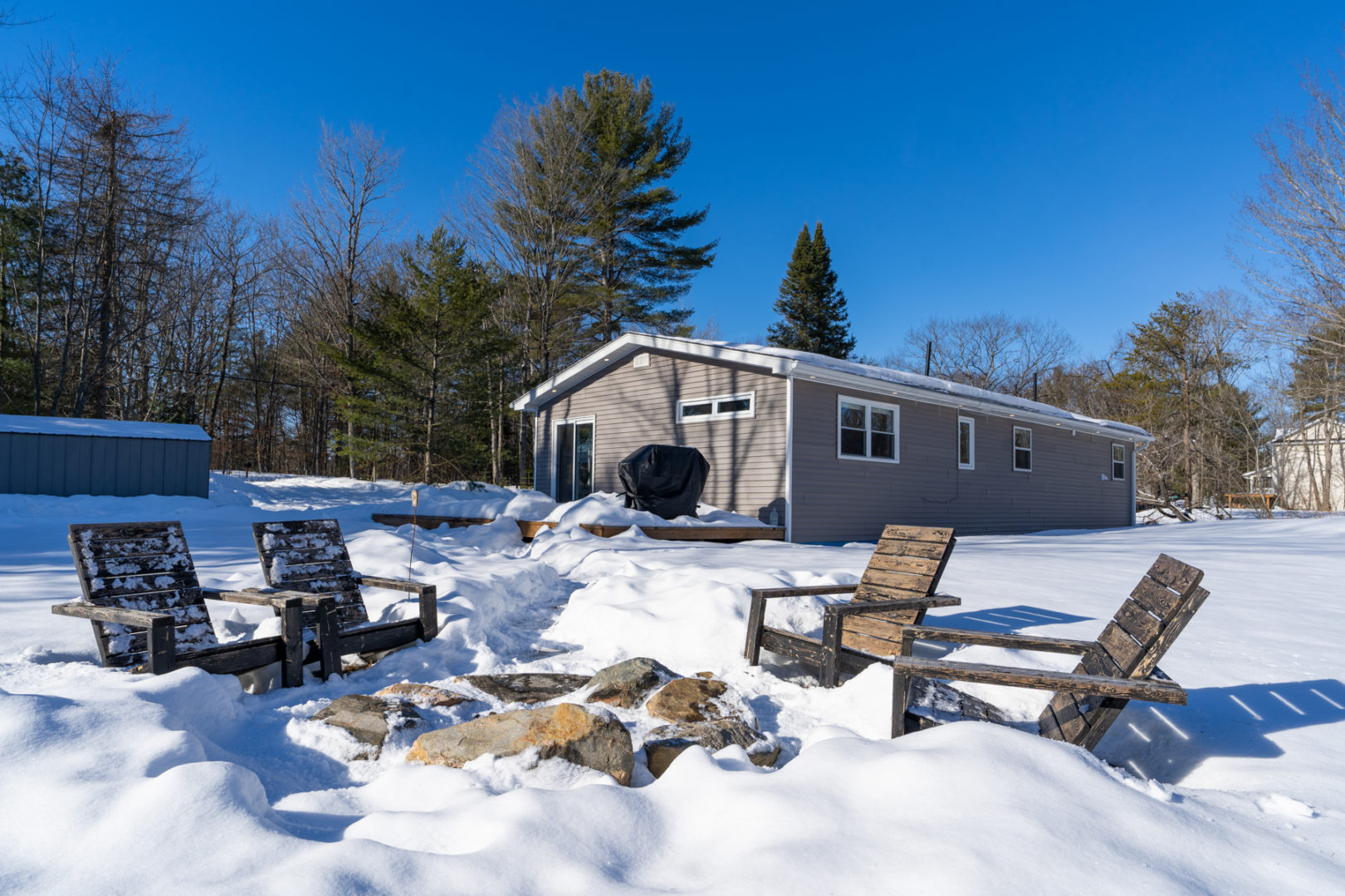 Exterior of a cottage bungalow, showing a large fire pit area and a big back deck extending off the side of the house. Surrounded by open snowy yard and trees.
