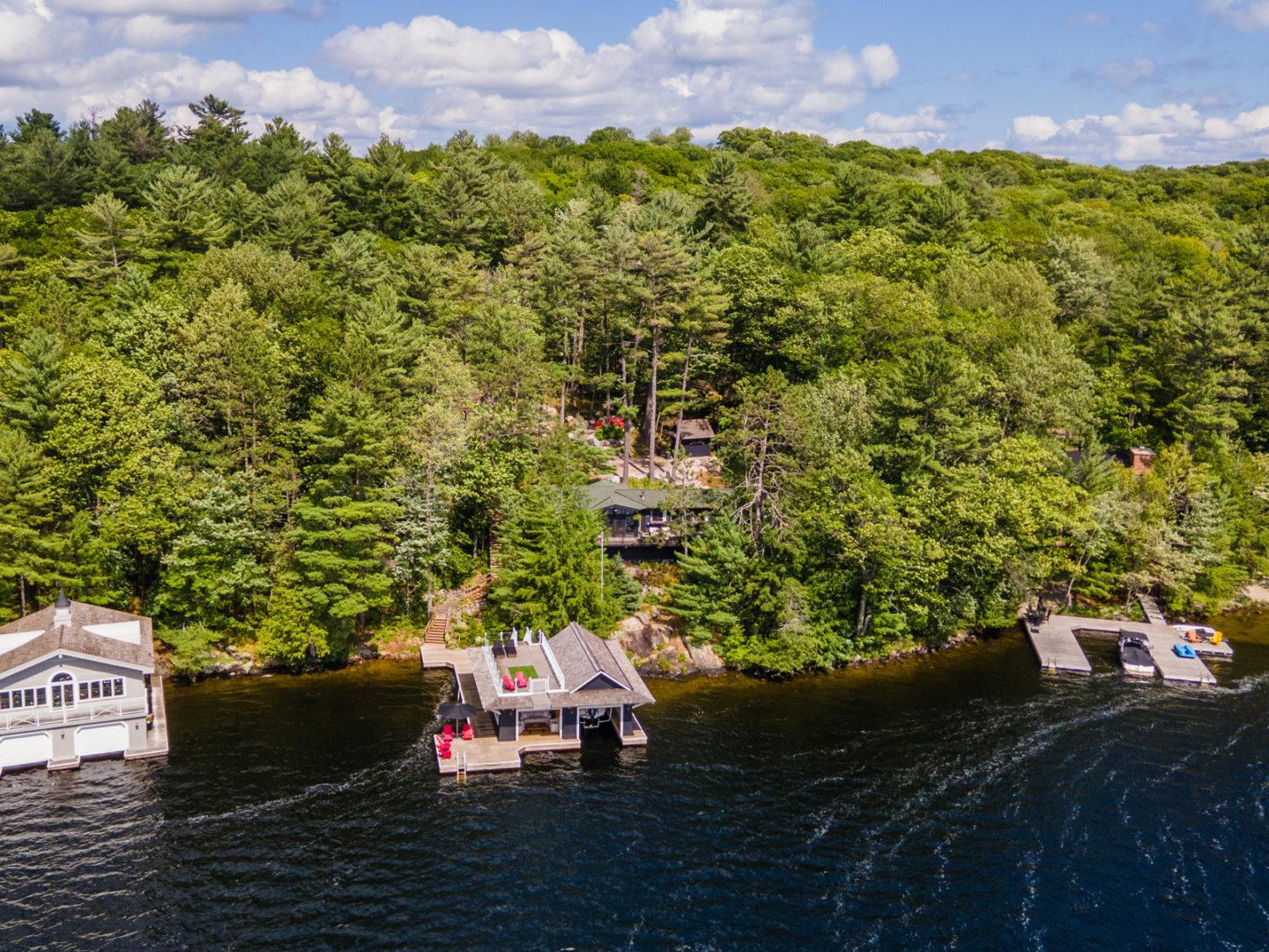 Shoreline of a cottage property, with a boathouse and a large dock in the water and a large cottage peeking through the trees in behind.