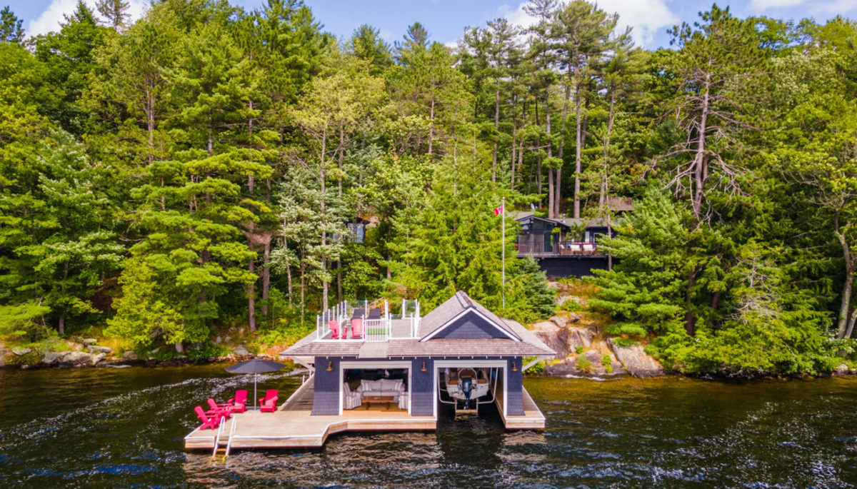 A boathouse with a large dock sits at the edge of a lake, with a cottage in behind the many trees on the shore.