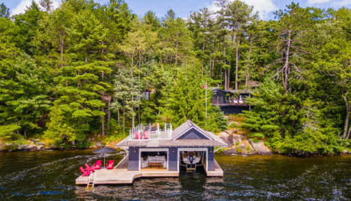 A boathouse with a large dock sits at the edge of a lake, with a cottage in behind the many trees on the shore.