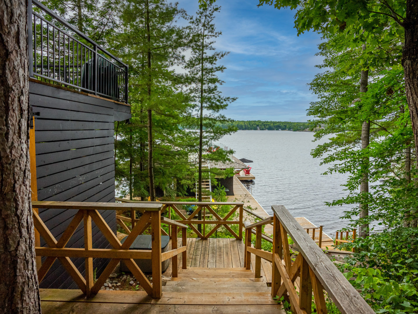 A wooden deck and stairway off a large cottage, looking out to a view of the lake through the trees.