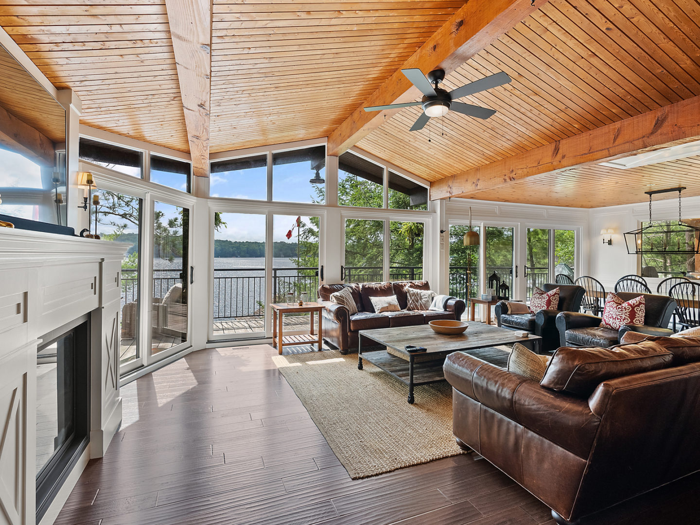 A bright, open-concept cottage living space with wood-panelled ceilings, floor-to-ceiling windows, and brown leather couches.