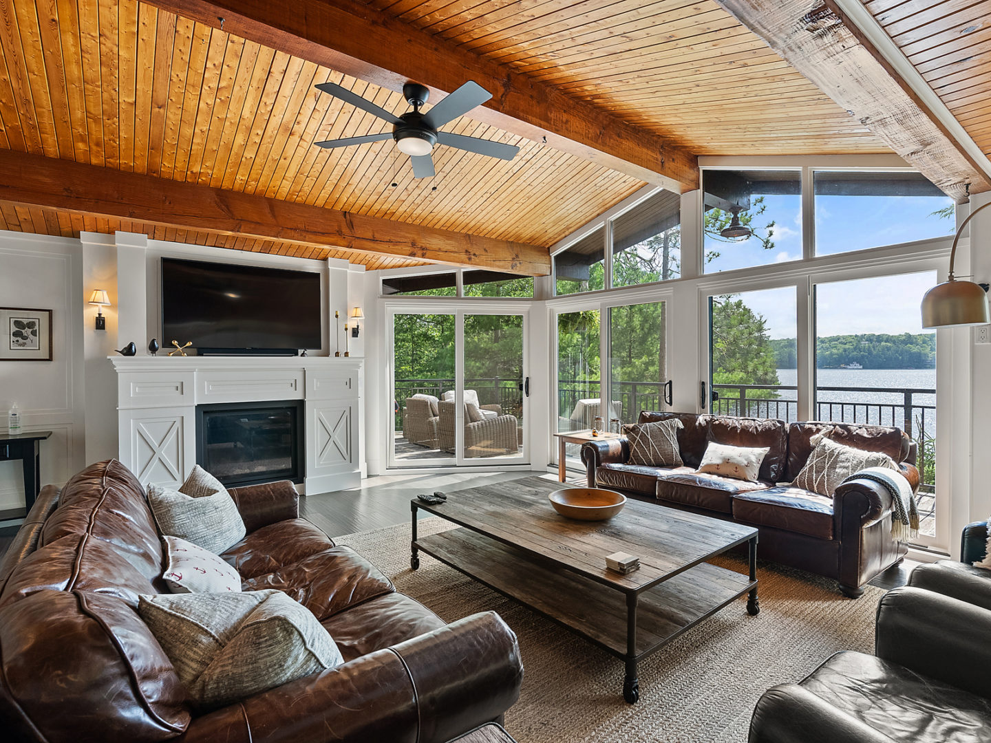 A luxury cottage living area with wood-panelled ceilings, floor-to-ceiling windows, brown leather couches, and a television..