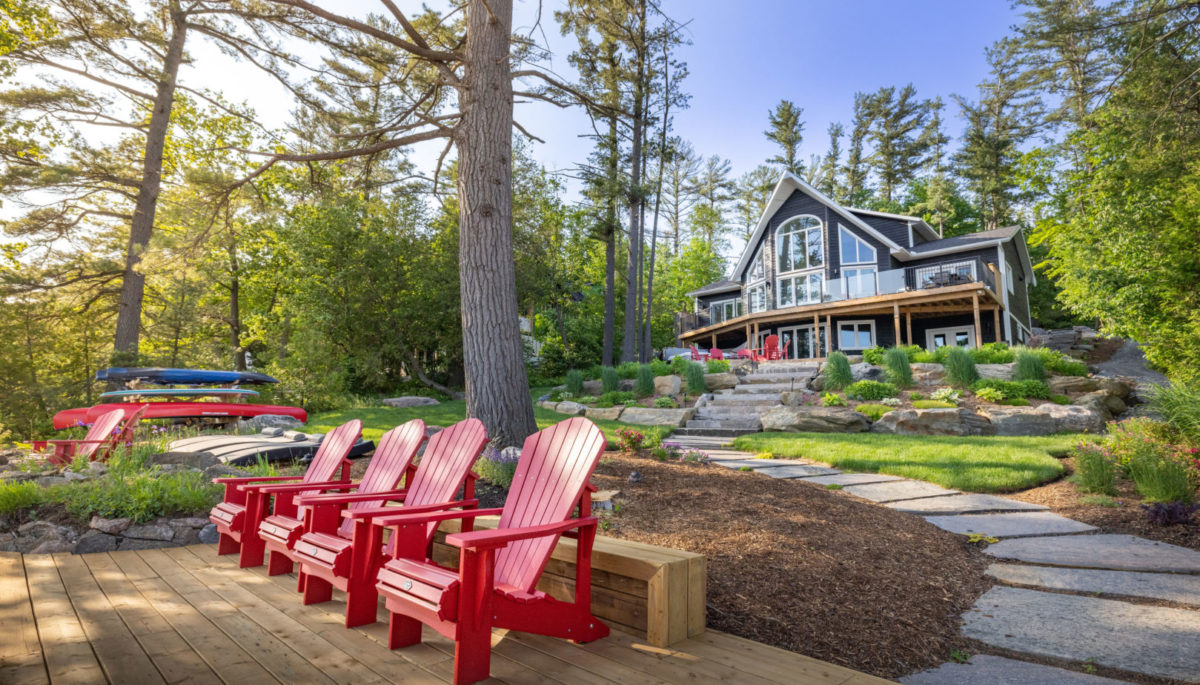 Four red Muskoka chairs sit on a dock. Landscaped stone steps lead up to a large cottage with huge windows and trees all around.