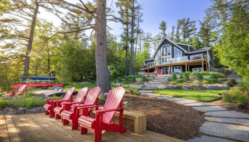 Four red Muskoka chairs sit on a dock. Landscaped stone steps lead up to a large cottage with huge windows and trees all around.