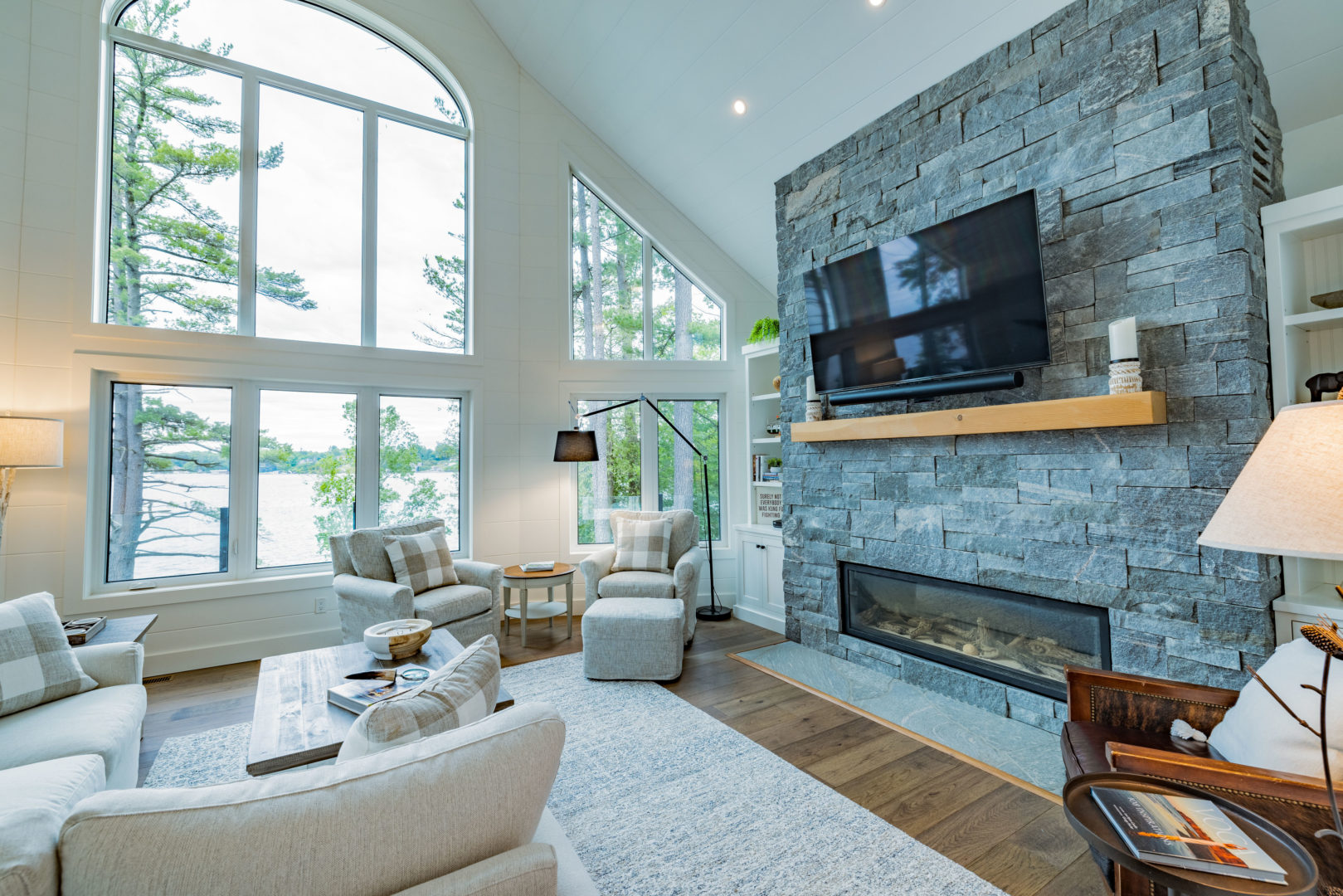A large living area with a couch, chairs, and a television mounted on a stone accent wall. Floor-to-ceiling windows provide a view of a large lake.