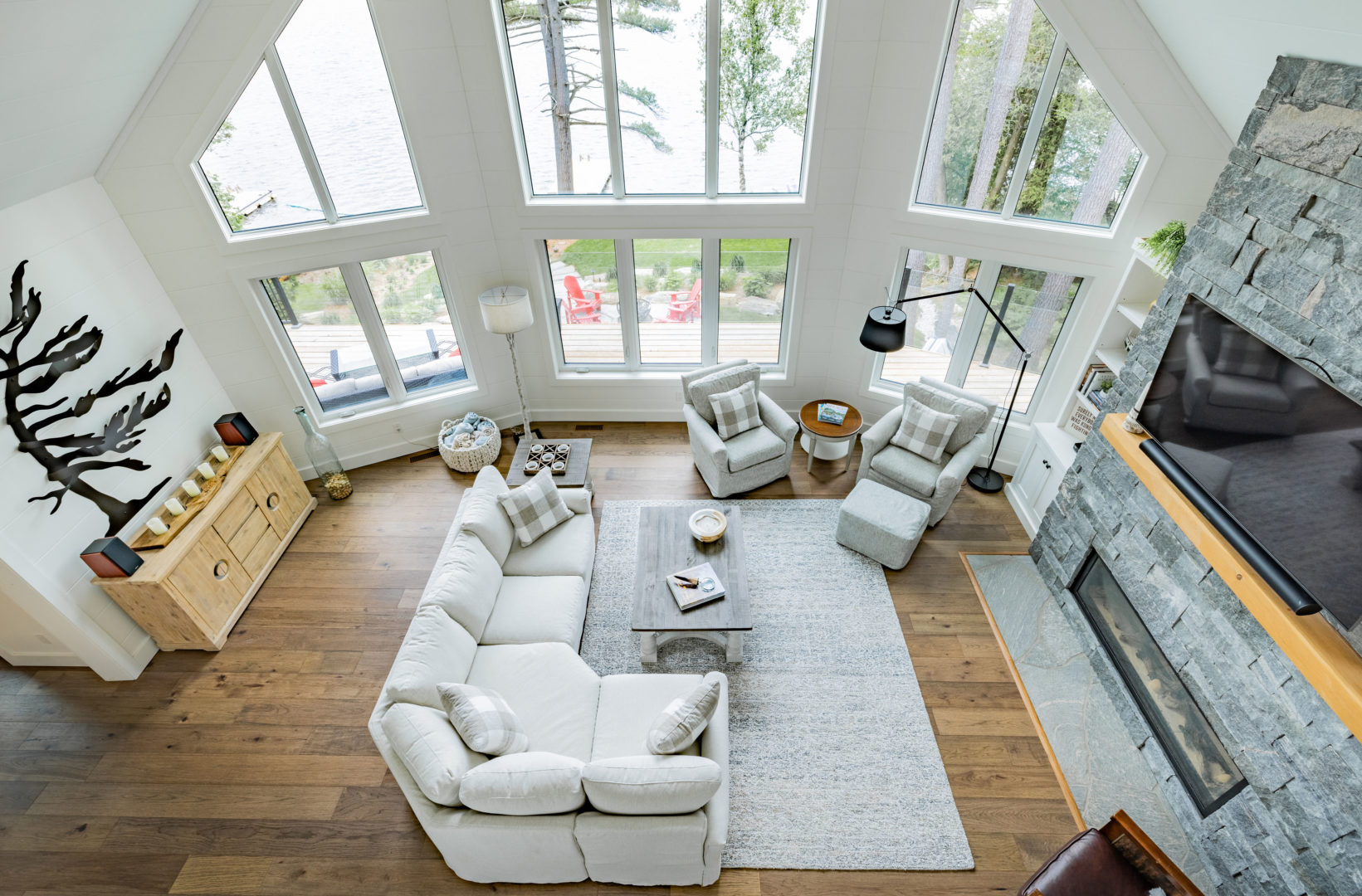 Overhead view of a large living area with a couch, chairs, and hardwood floors. Floor-to-ceiling windows provide a view of a large lake.