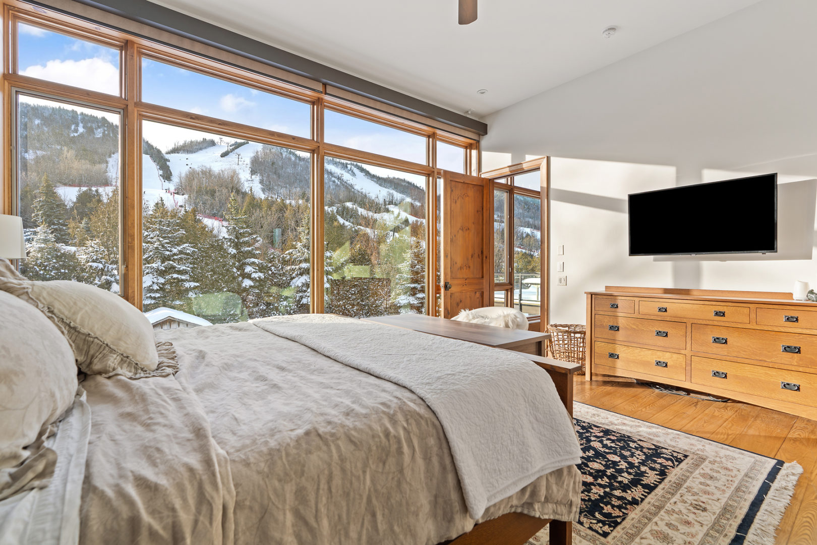 A large bedroom with a huge window across one wall. The view looks out on the snowy slope of a mountain.