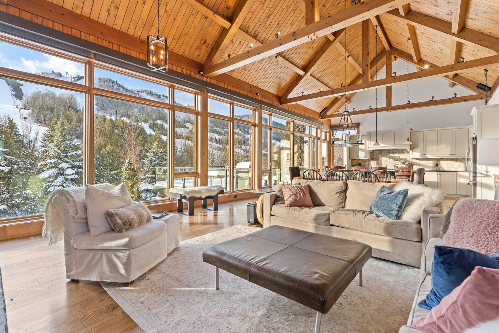 A huge open-concept space with a living and dining area. Wooden beams accent the high ceiling, and windows with a mountain view stretch all along one side of the house.