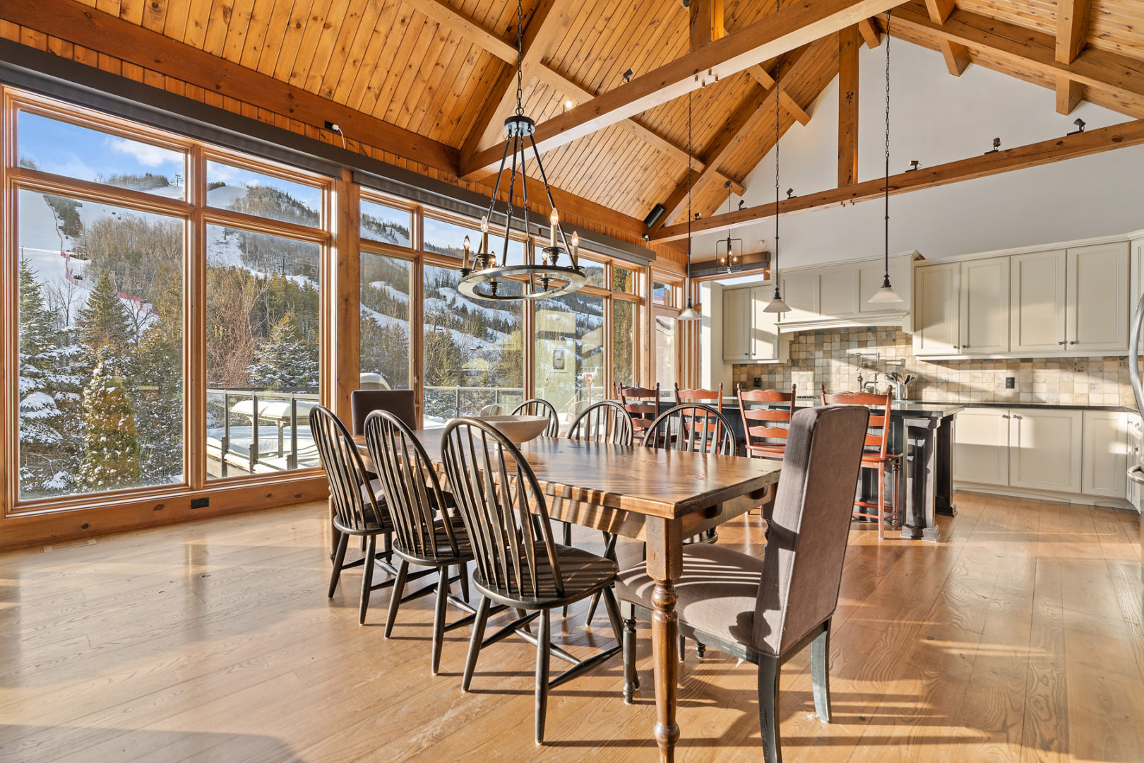 A dining area with a table and chairs in the middle of a large open-concept space, leading into a kitchen. Wooden beams accent the high ceiling, and windows with a mountain view stretch all along one side of the house.