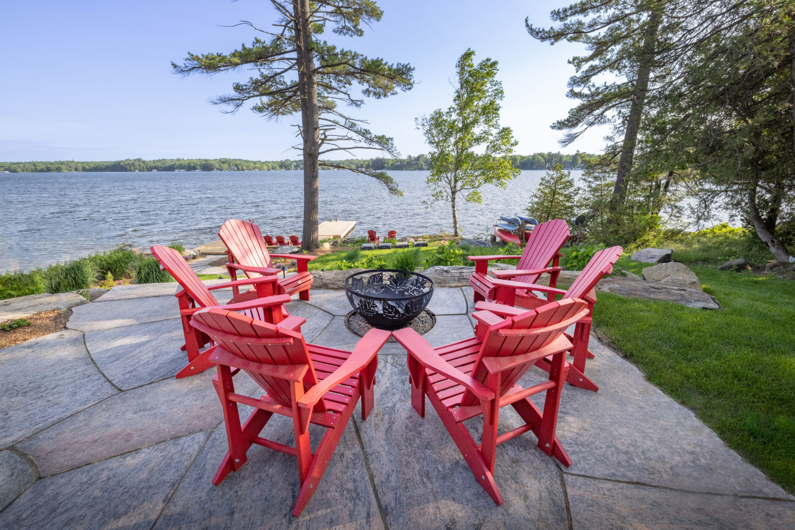 A fire pit surrounded by red Muskoka chairs sits on a patio overlooking a large lake.