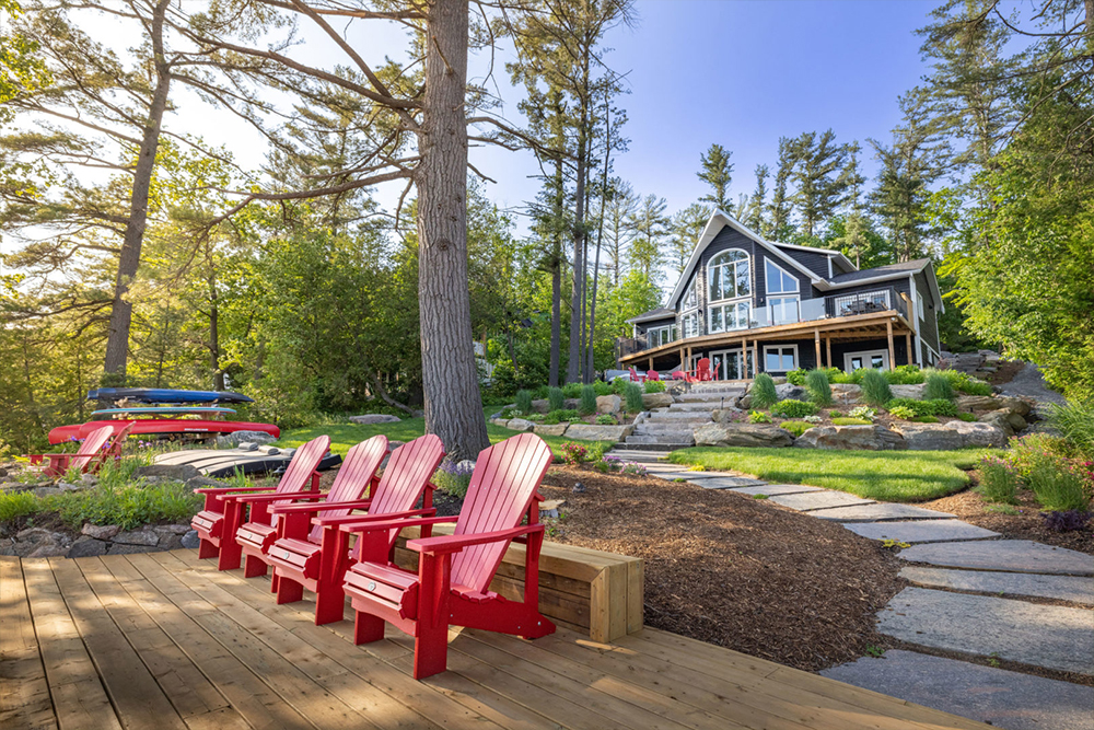 Four red Muskoka chairs sit on a dock. Landscaped stone steps lead up to a large cottage with huge windows and trees all around.