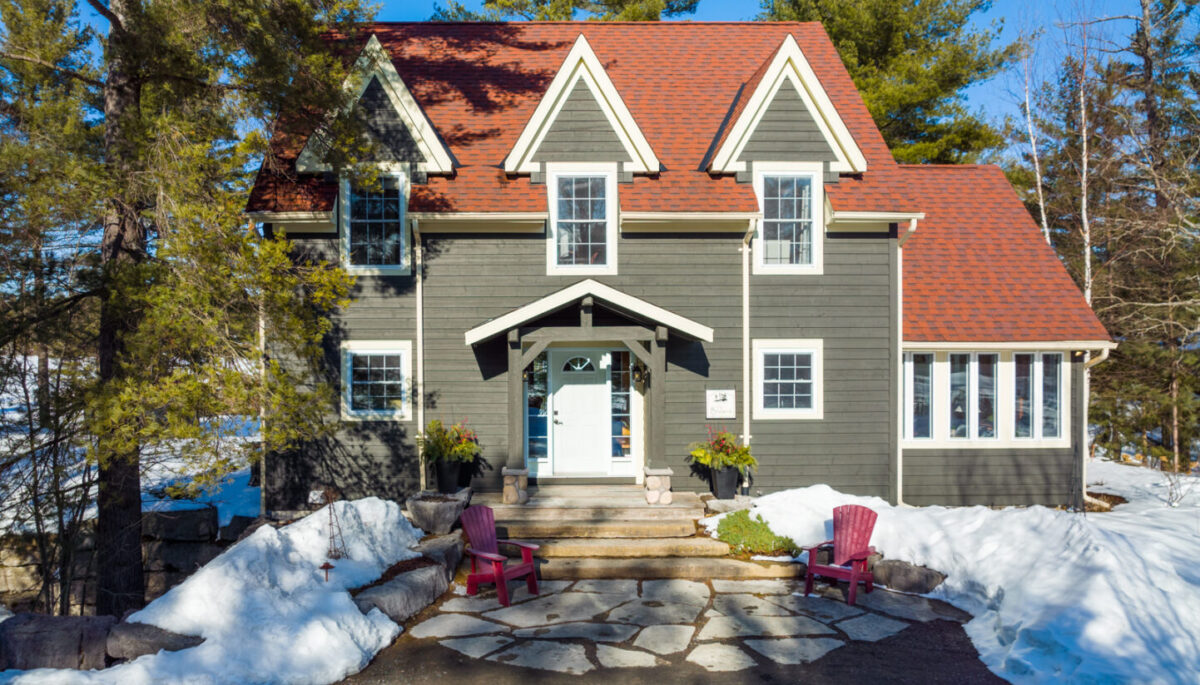 The front of a large lakefront home with a greenish grey exterior and a red roof.