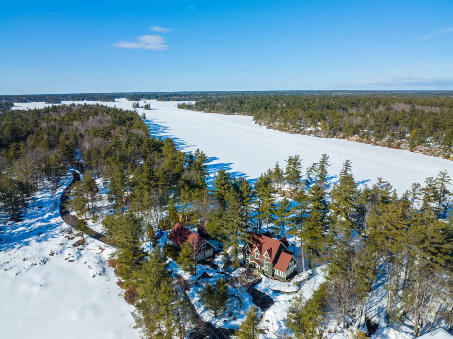 Overhead view of a large lakefront property in the wintertime. A large cottage with a red roof sits on a snow-covered waterfront surrounded by evergreen trees.