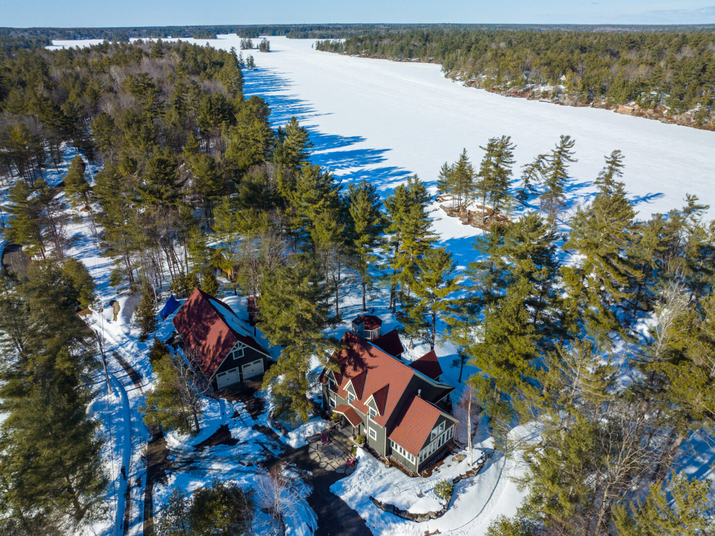 Overhead view of a large lakefront property in the wintertime. A large cottage with a red roof sits on a snow-covered waterfront surrounded by evergreen trees.