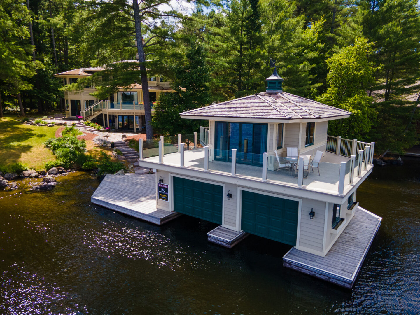 A large, two-storey boathouse sitting on a dock. Stone steps in behind lead to a large cottage.
