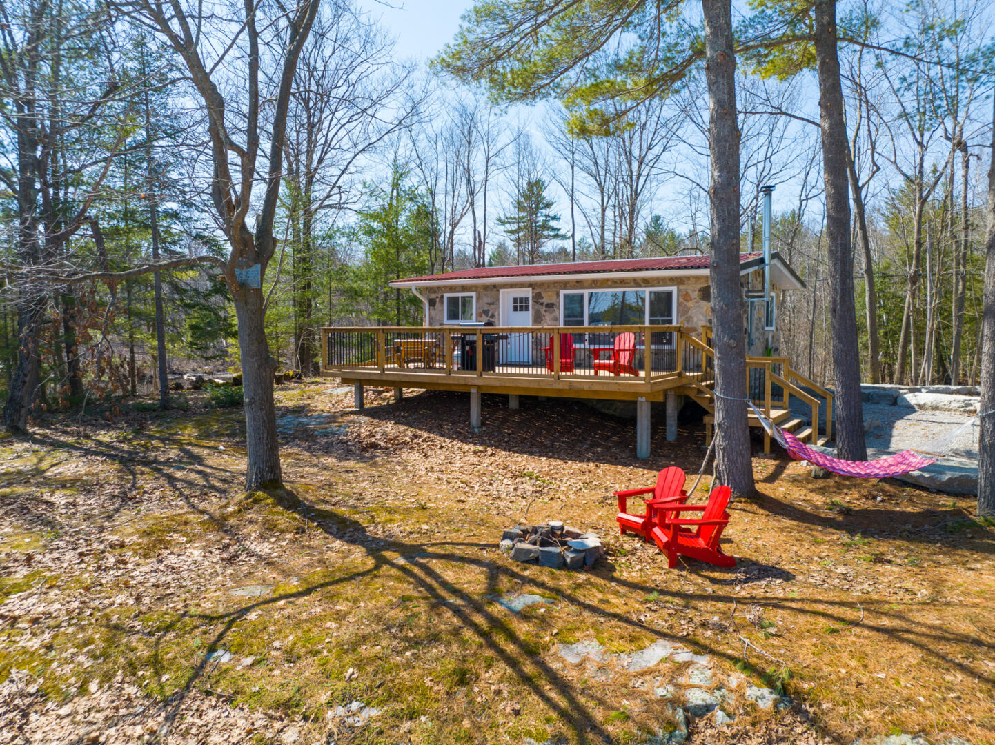 Outdoor space of a small cottage that has large windows and a big back deck. There are trees all around, and two red Muskoka chairs sit in front of a small firepit.