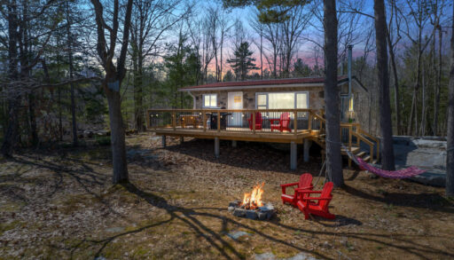 Outdoor space of a small cottage that has large, lit-up windows and a big back deck. There are trees all around, and two red Muskoka chairs sit in front of a small lit firepit.