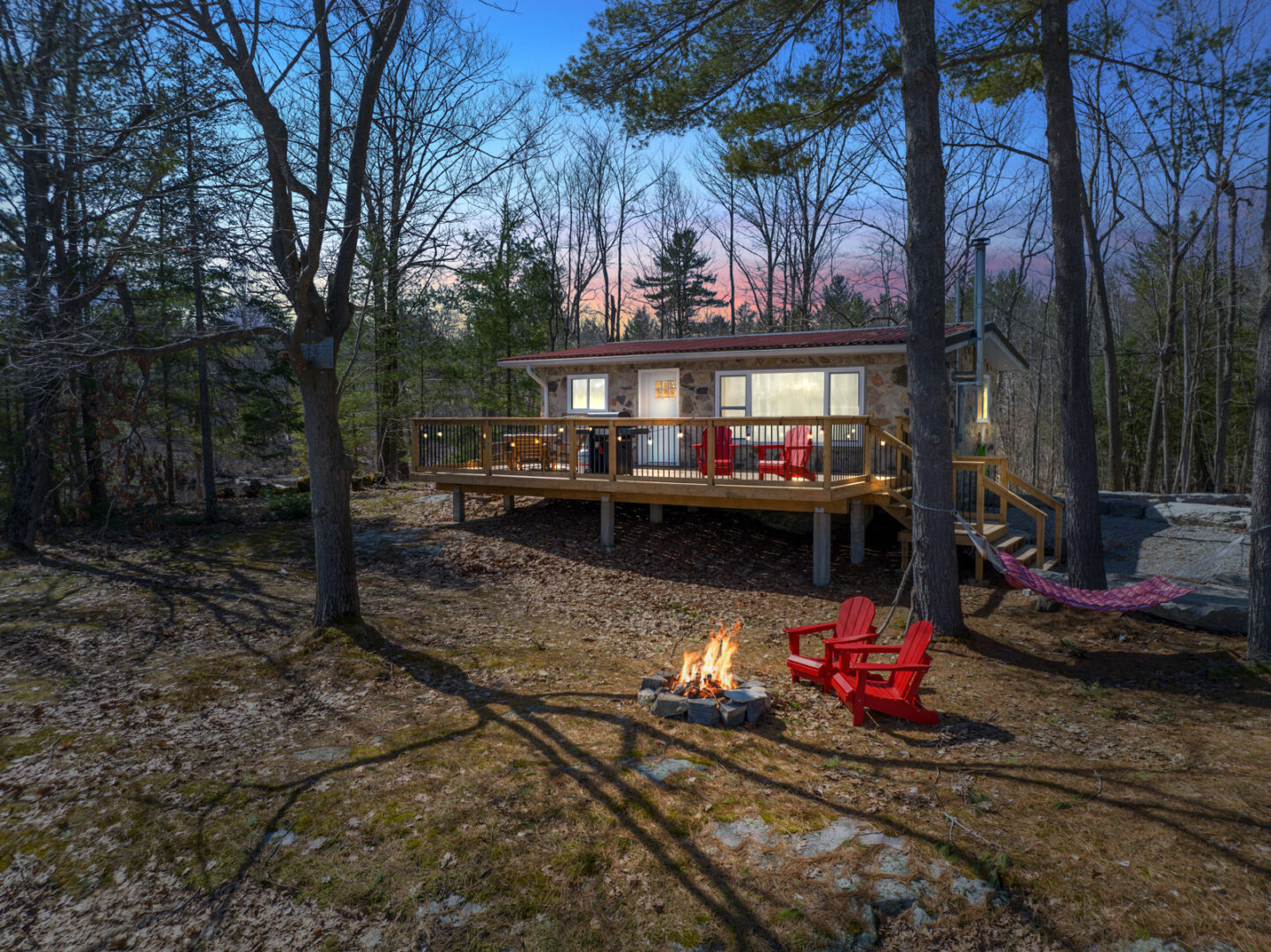 Outdoor space of a small cottage that has large, lit-up windows and a big back deck. There are trees all around, and two red Muskoka chairs sit in front of a small lit firepit.