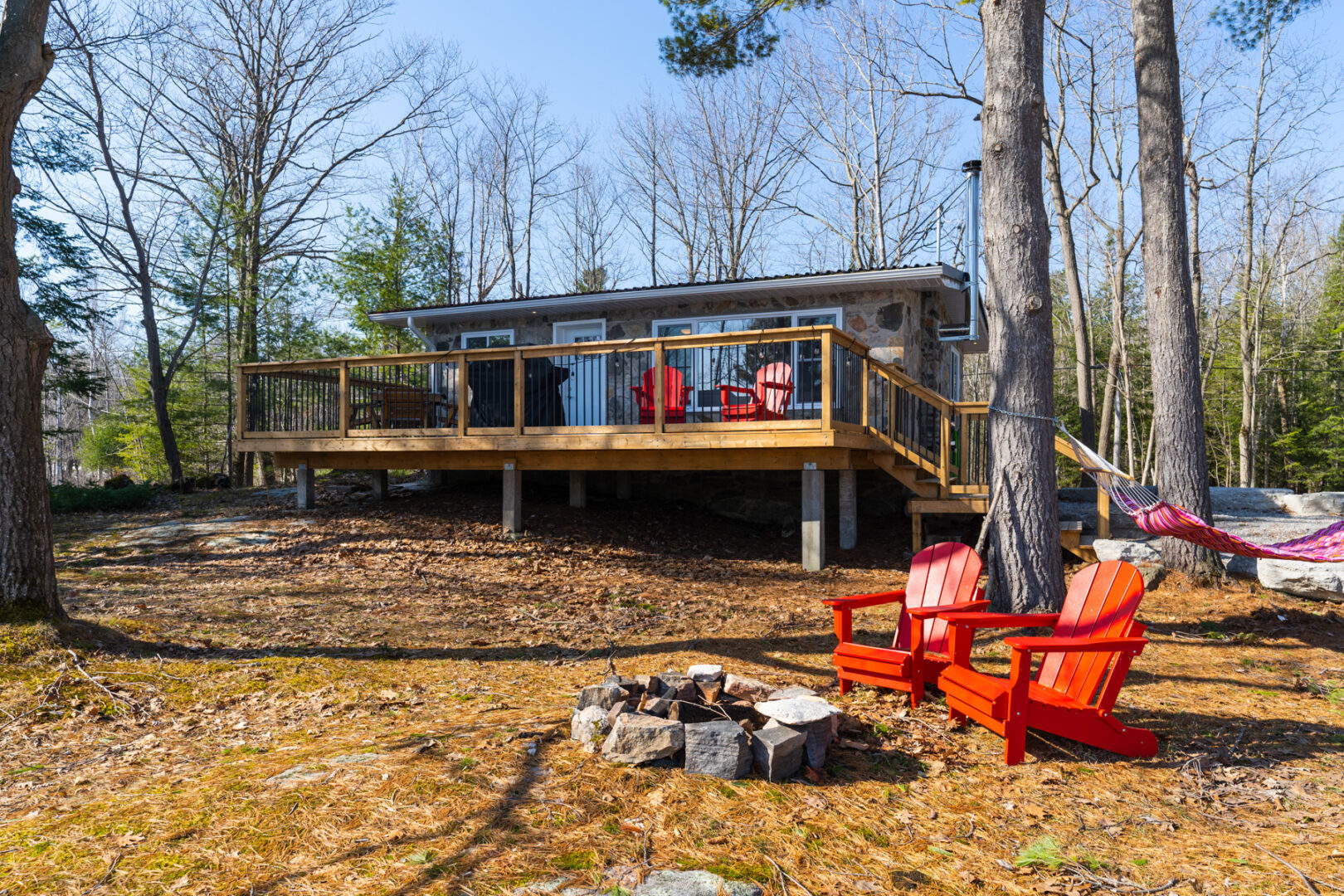 A small cottage with a deck and lots of windows sits on a large open yard surrounded by trees.