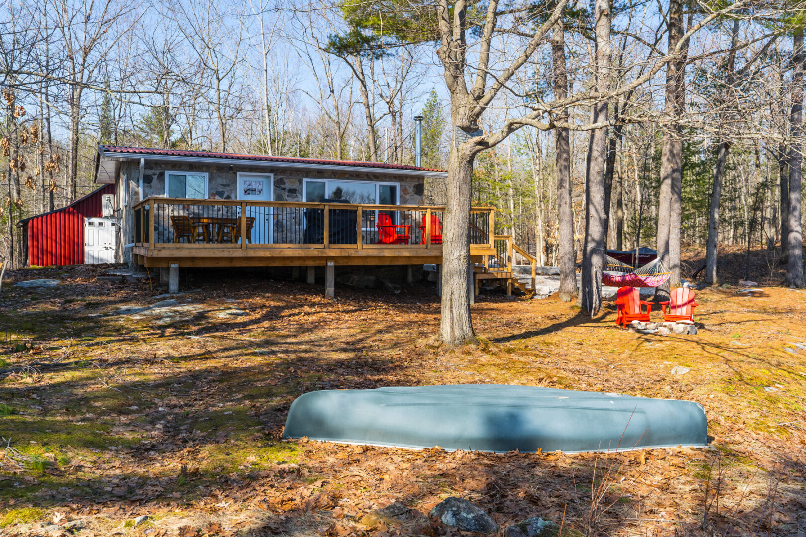 Outdoor space around a small cottage that has lots of windows and a big back deck. An upside-down canoe lies on the ground, there are trees all around, and Muskoka chairs sit in front of a small firepit off to the side.