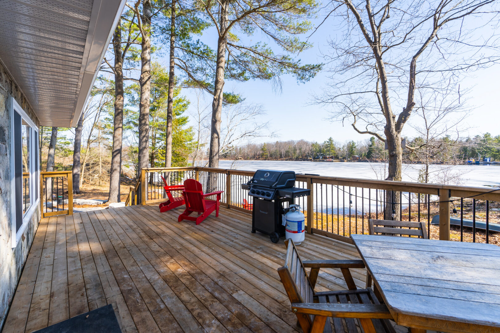 A large wooden deck off the back of a small cottage, with a barbecue, Muskoka chairs, and a view of the lake.