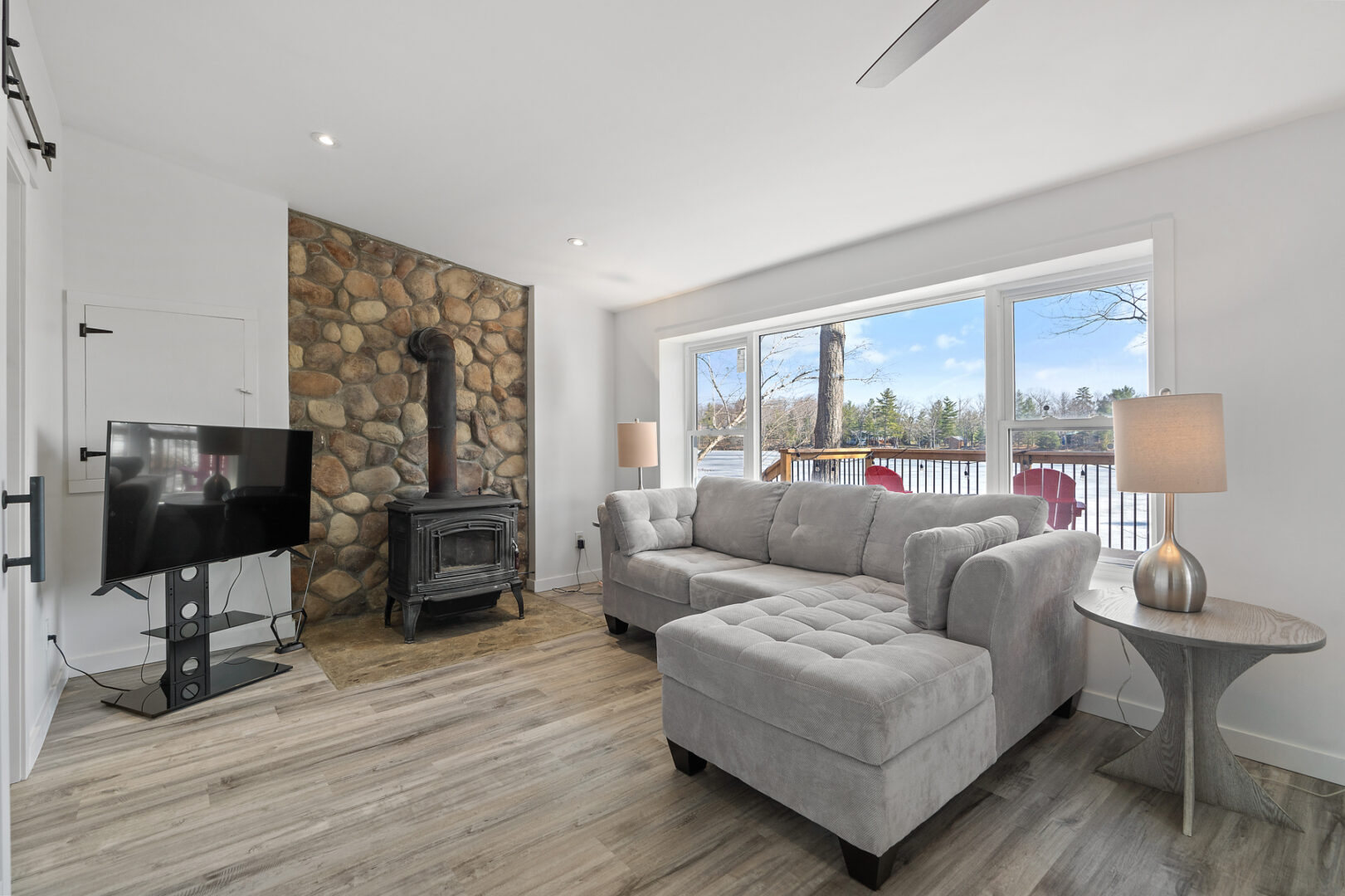 A small, bright cottage living space with a wood-burning fireplace, a sectional couch, hardwood flooring and large windows with a view of the lake.