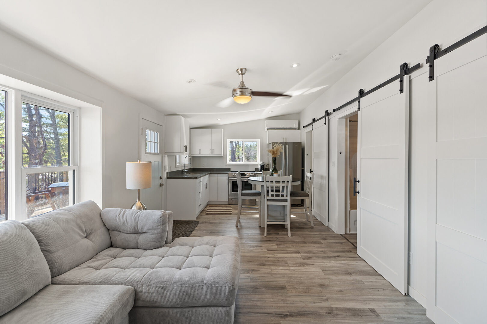 A small, open-concept cottage living space with hardwood flooring, a sectional couch, a small dining table and a modern kitchen.