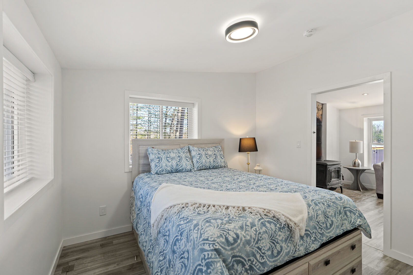 A bright, modest bedroom with a large bed, two windows, and a door leading out to a living room area.