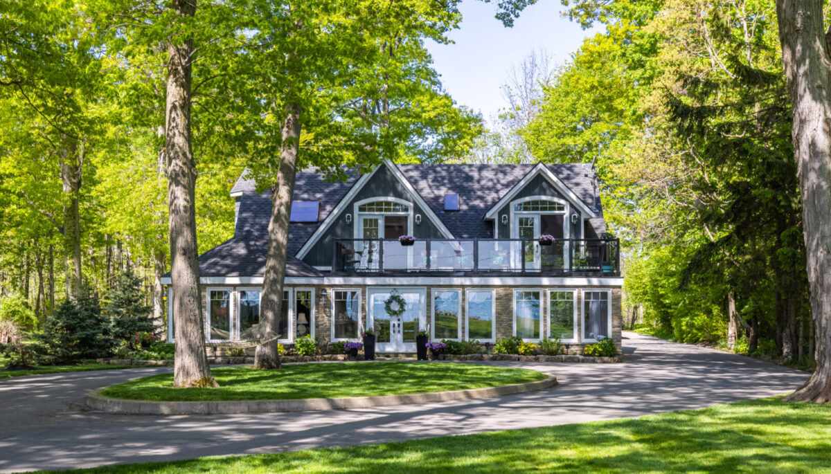 A large, lake-house style cottage with lots of windows sits on a manicured grass lot, surrounded by trees.