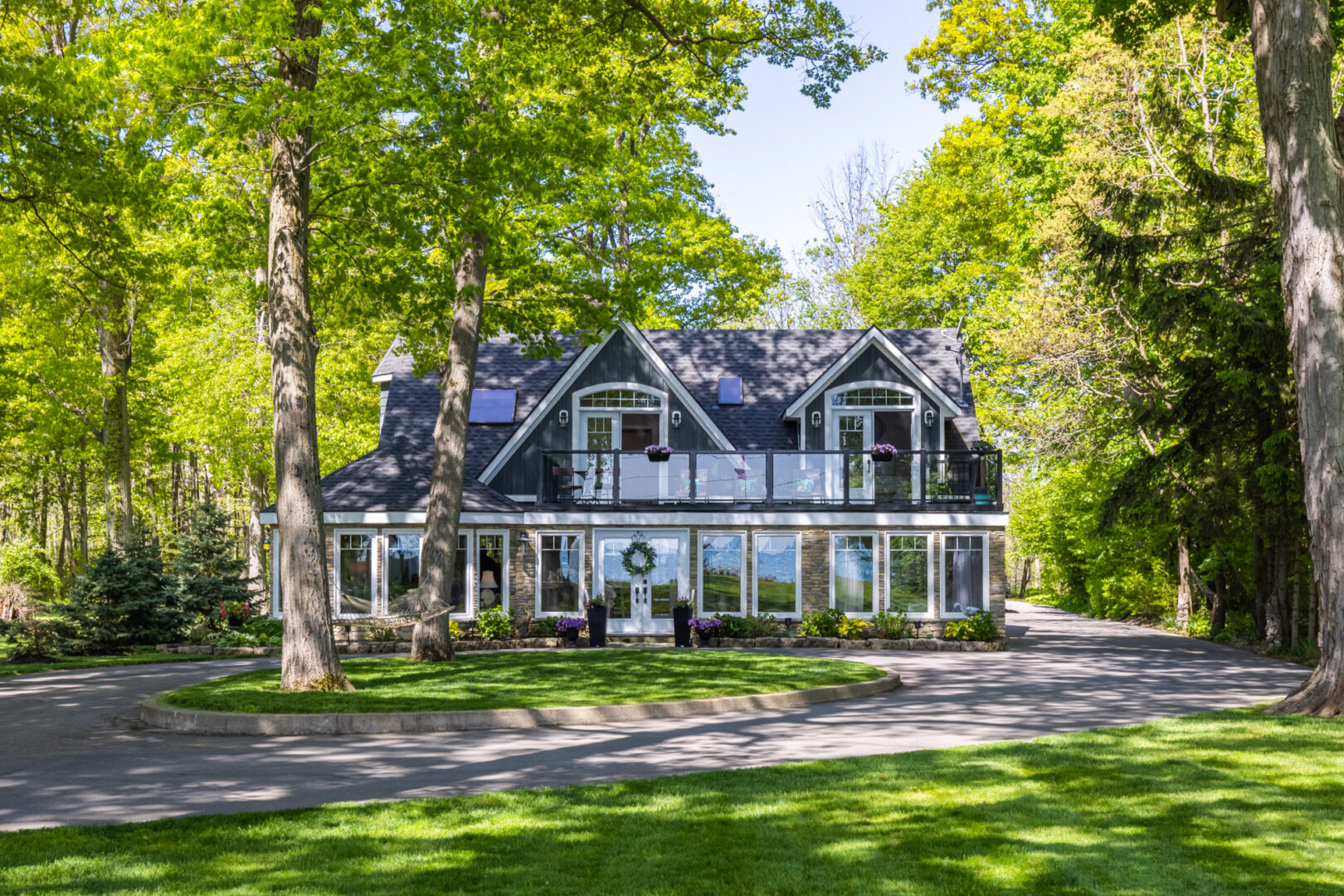 A large, lake-house style cottage with lots of windows sits on a manicured grass lot, surrounded by trees.