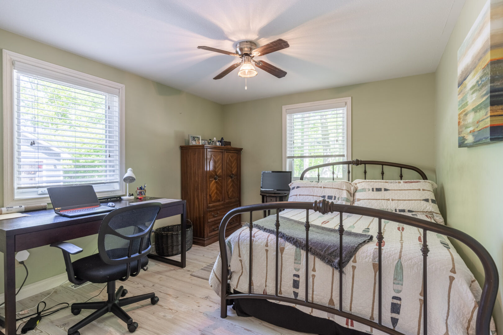 A small, bright bedroom with a large bed, a desk, a small window, and a ceiling fan.