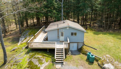 A small cottage with a large wrap-around deck sits in the centre of a clearing surrounded by trees.